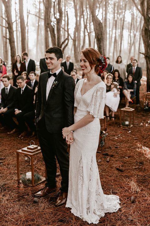 Elegant woodland wedding ceremony with long white dress and black tuxedo in a misty forest setting.