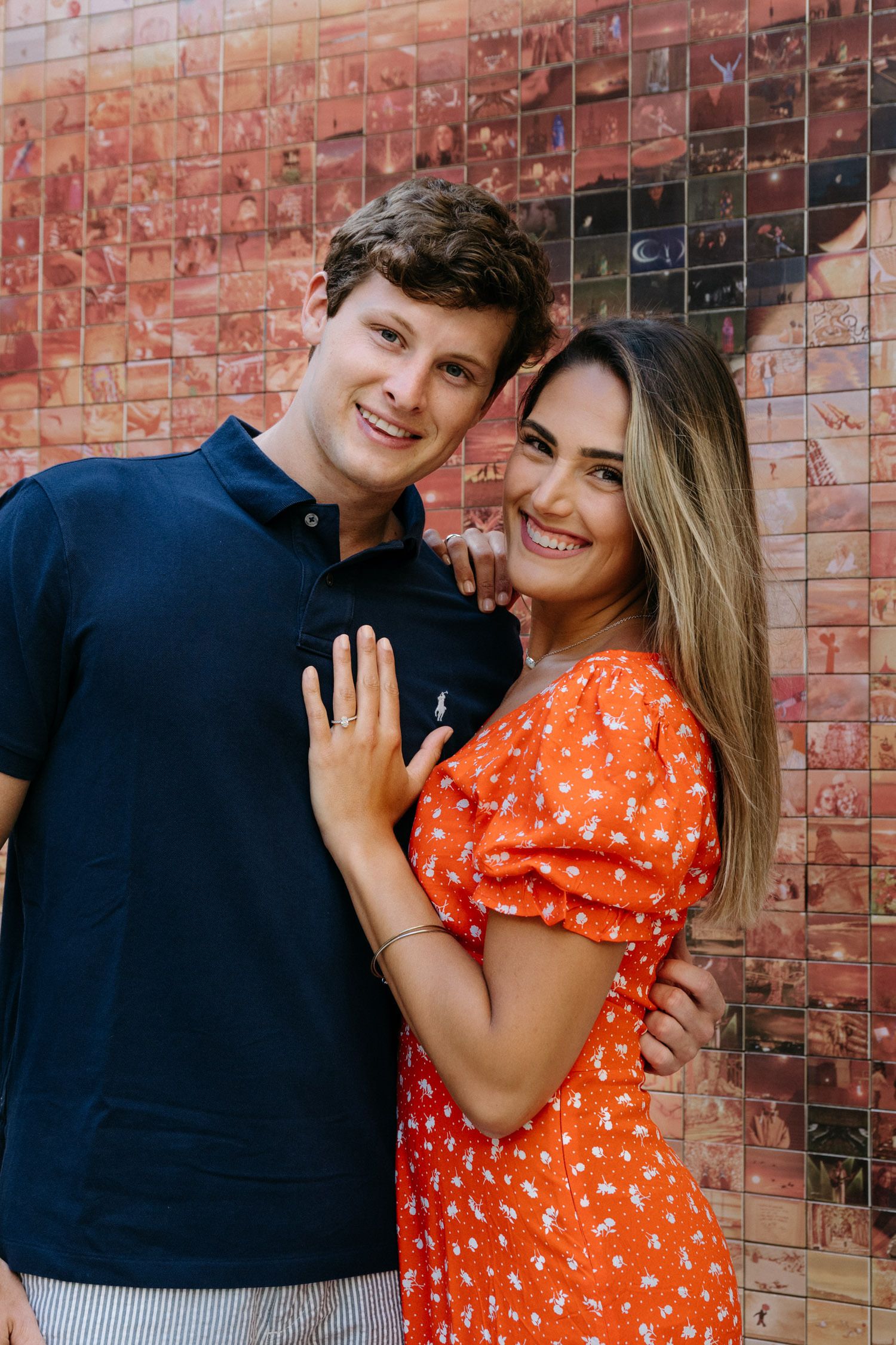 Couple in navy and orange outfits posing playfully against brick wall.
