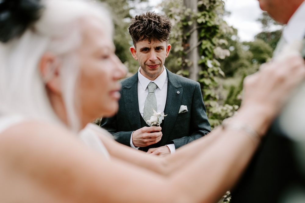 A wedding ceremony showing the groom in a gray suit with a silver tie reading from a card during the outdoor vows.