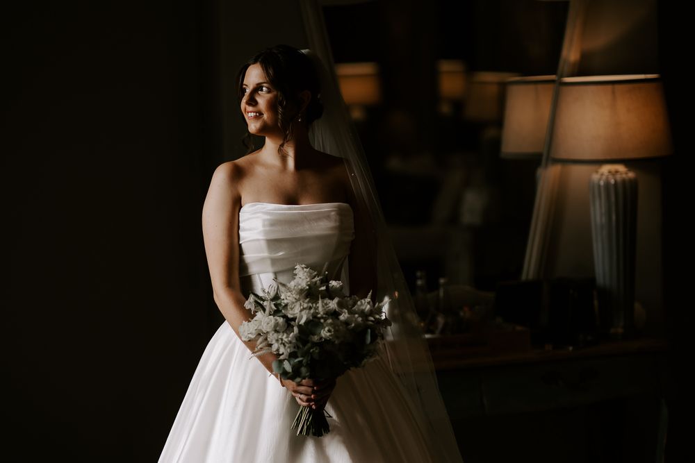 A bride in a strapless white gown holds a delicate bouquet while standing in moody, dramatic lighting.