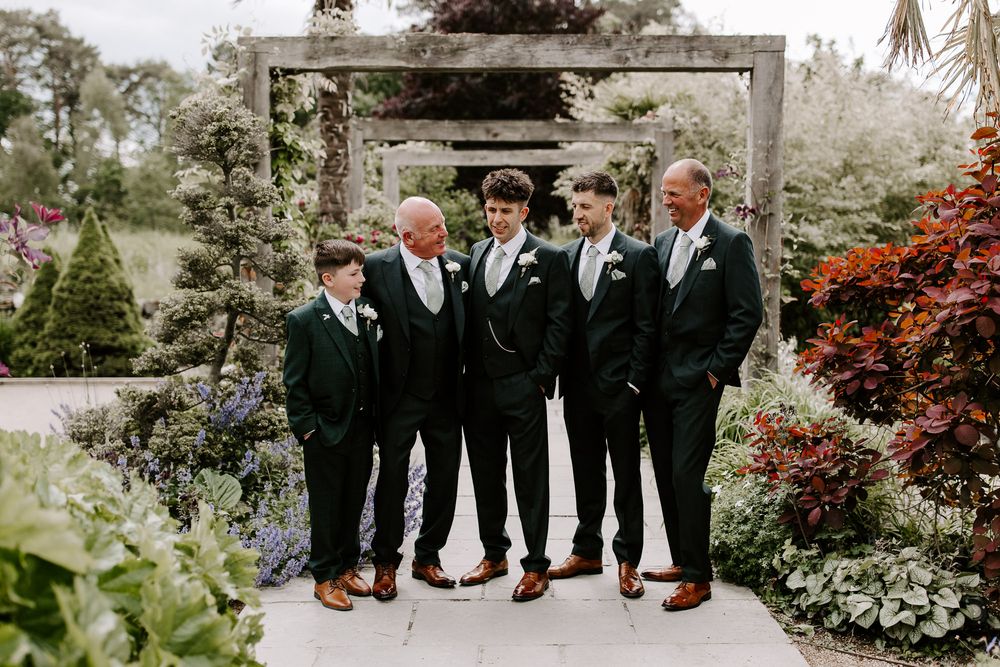 A group in formal black suits and brown shoes pose together in a garden archway surrounded by blooming flowers.