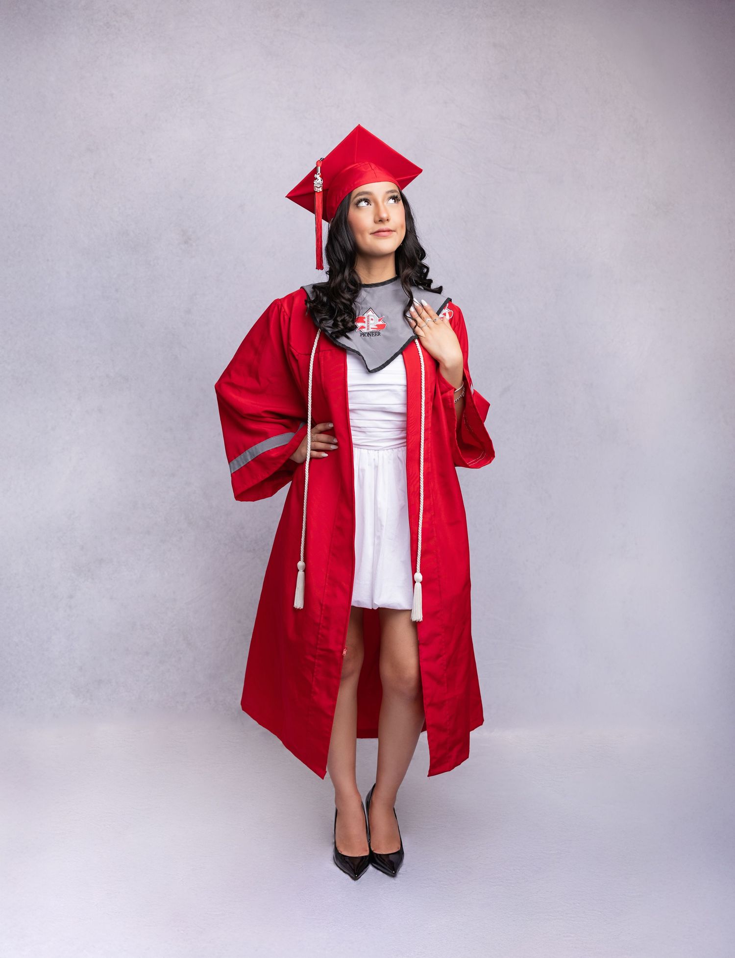 Graduate wearing traditional red cap and gown with white dress and black heels poses against gray background.
