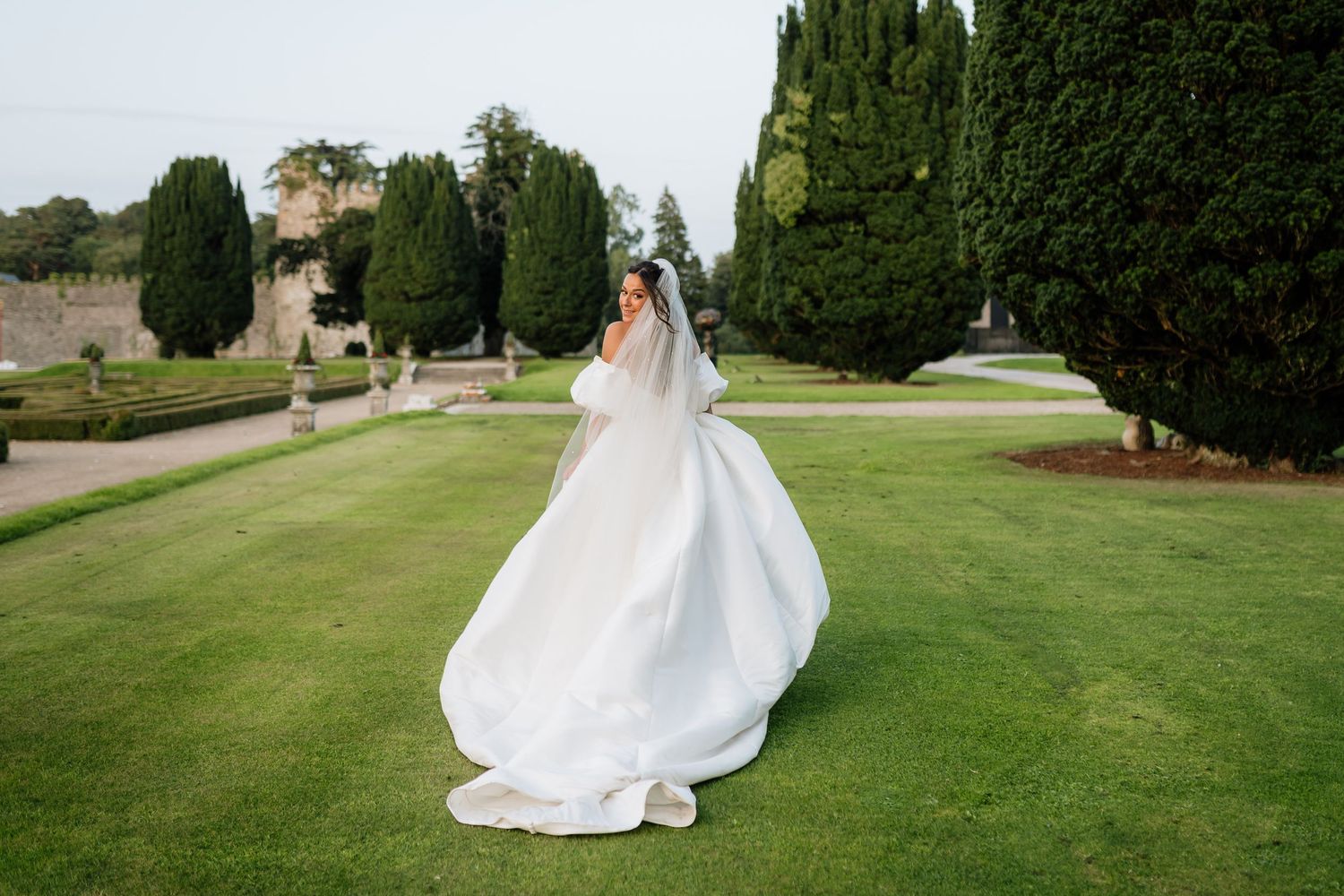 A flowing white wedding dress swirls on manicured garden grounds with tall cypress trees and hedges in the background.