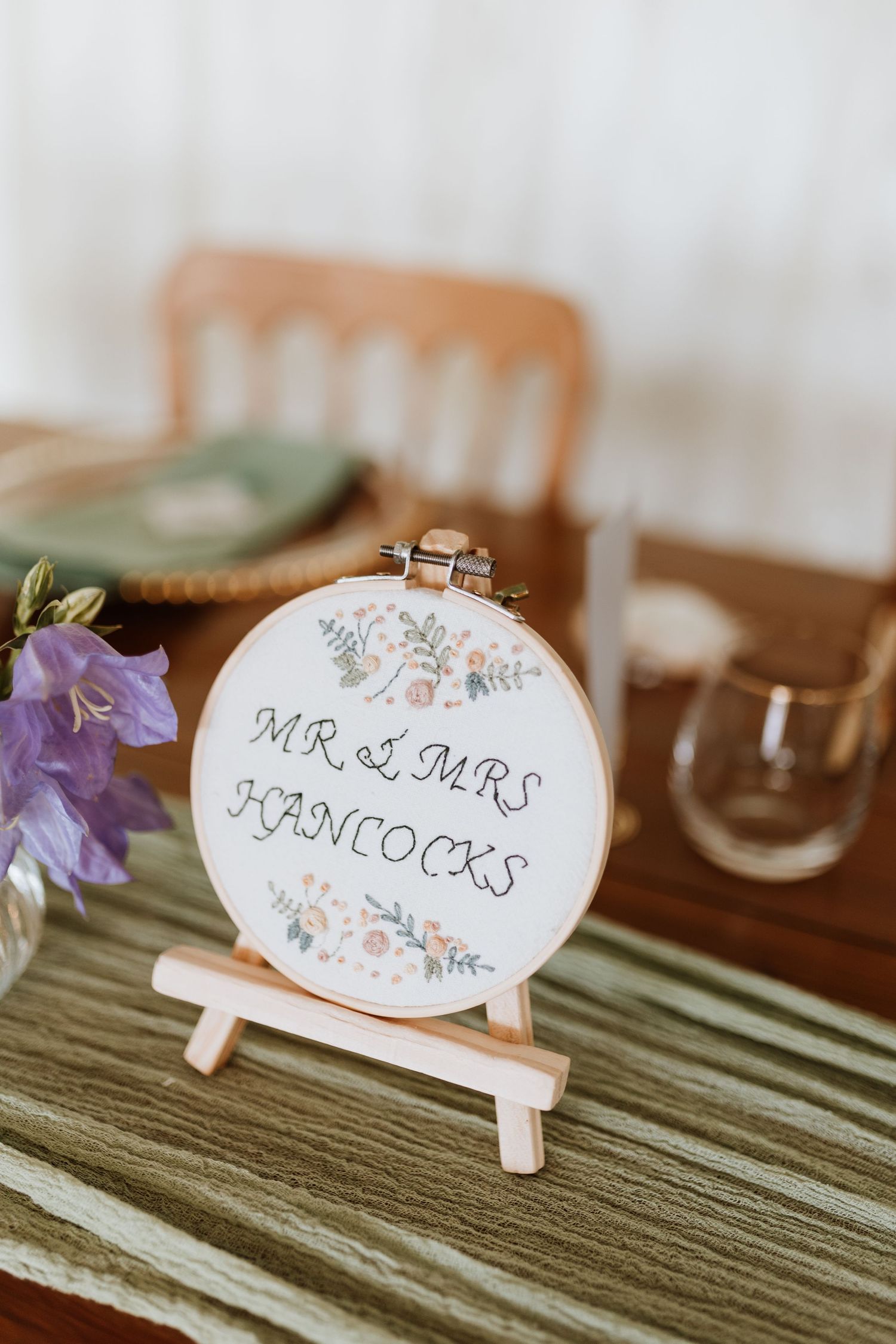 Custom embroidered hoop art displaying 'Mr & Mrs Hancocks' with floral details on a wooden stand surrounded by decor.