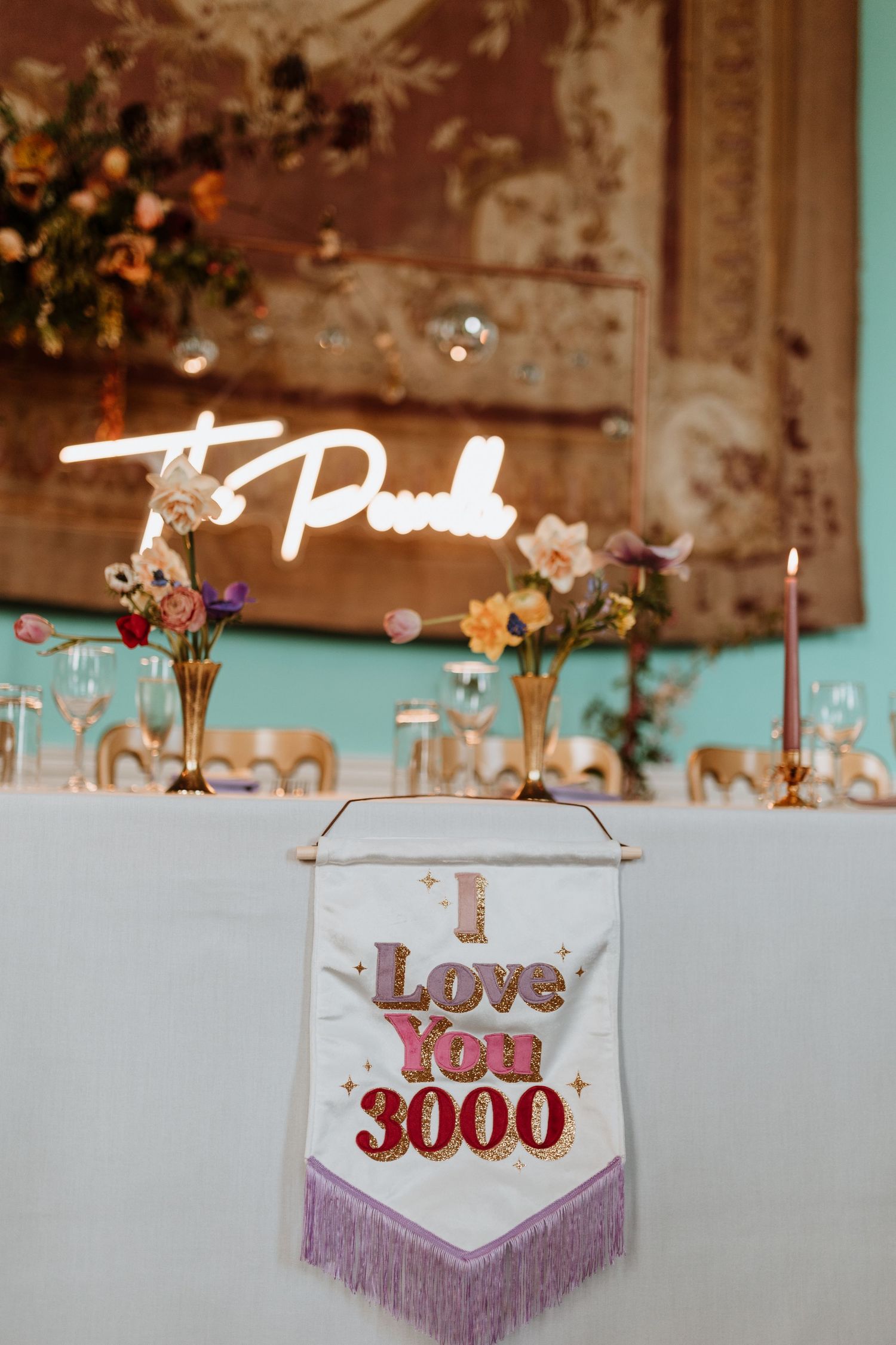 A decorative 'I Love You 3000' banner with pink fringe hangs beneath a neon sign with floral arrangements in a wedding setting.