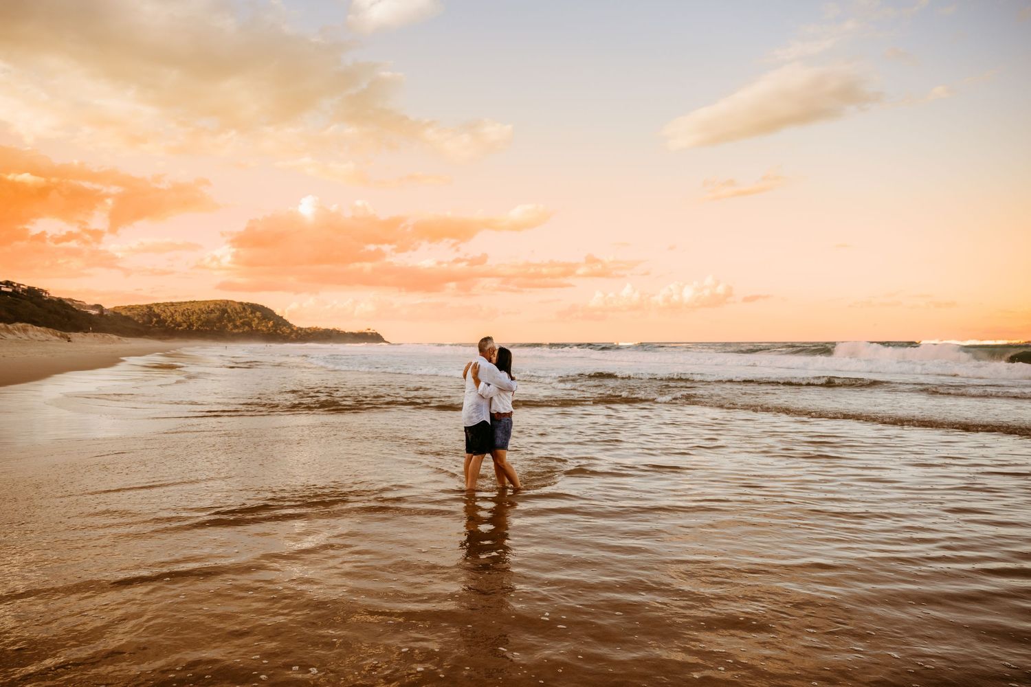 A couple stands in shallow ocean waves during a golden sunset at a scenic beach.
