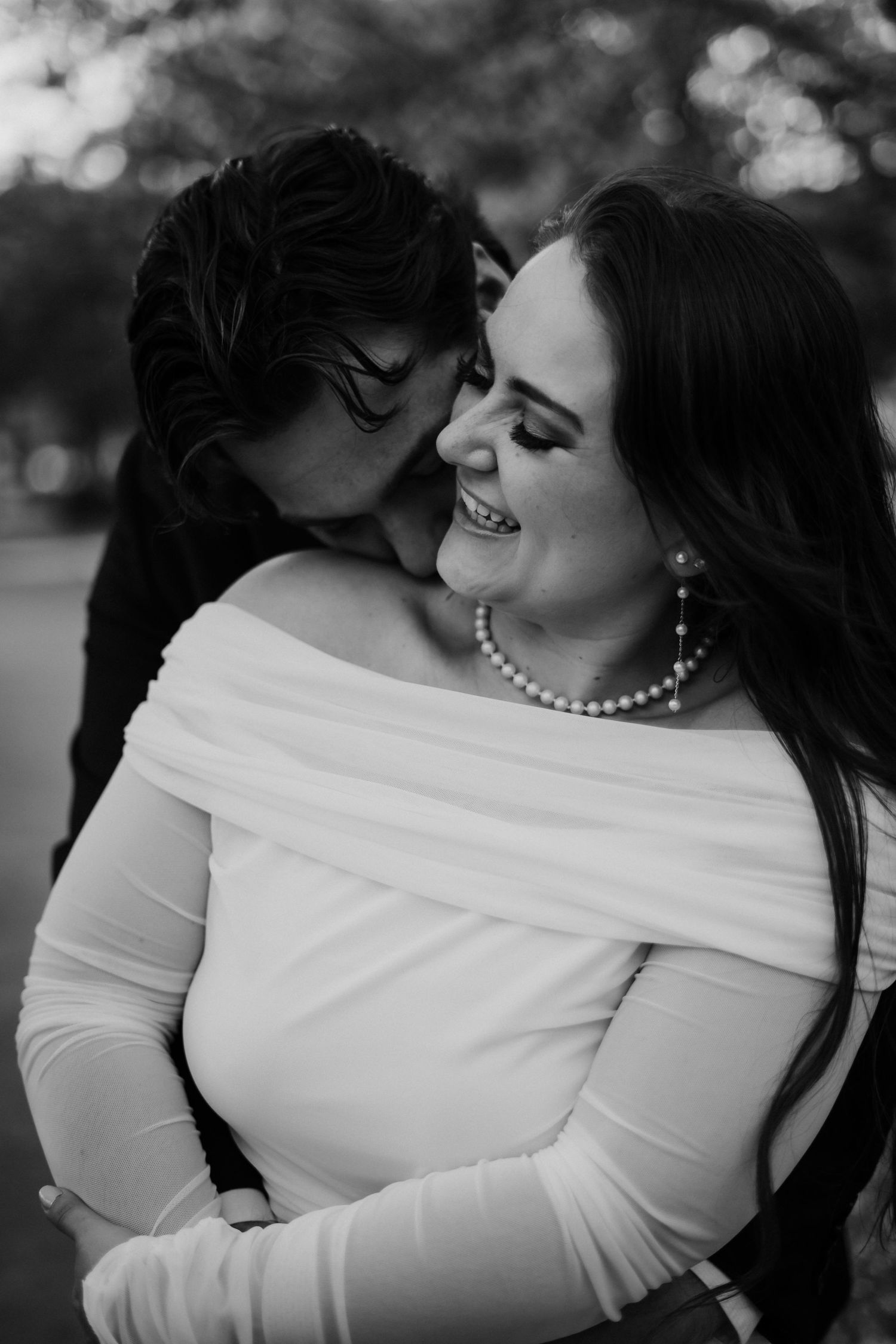 Black and white portrait of an intimate romantic moment showing affection while wearing white dress and pearl necklace.