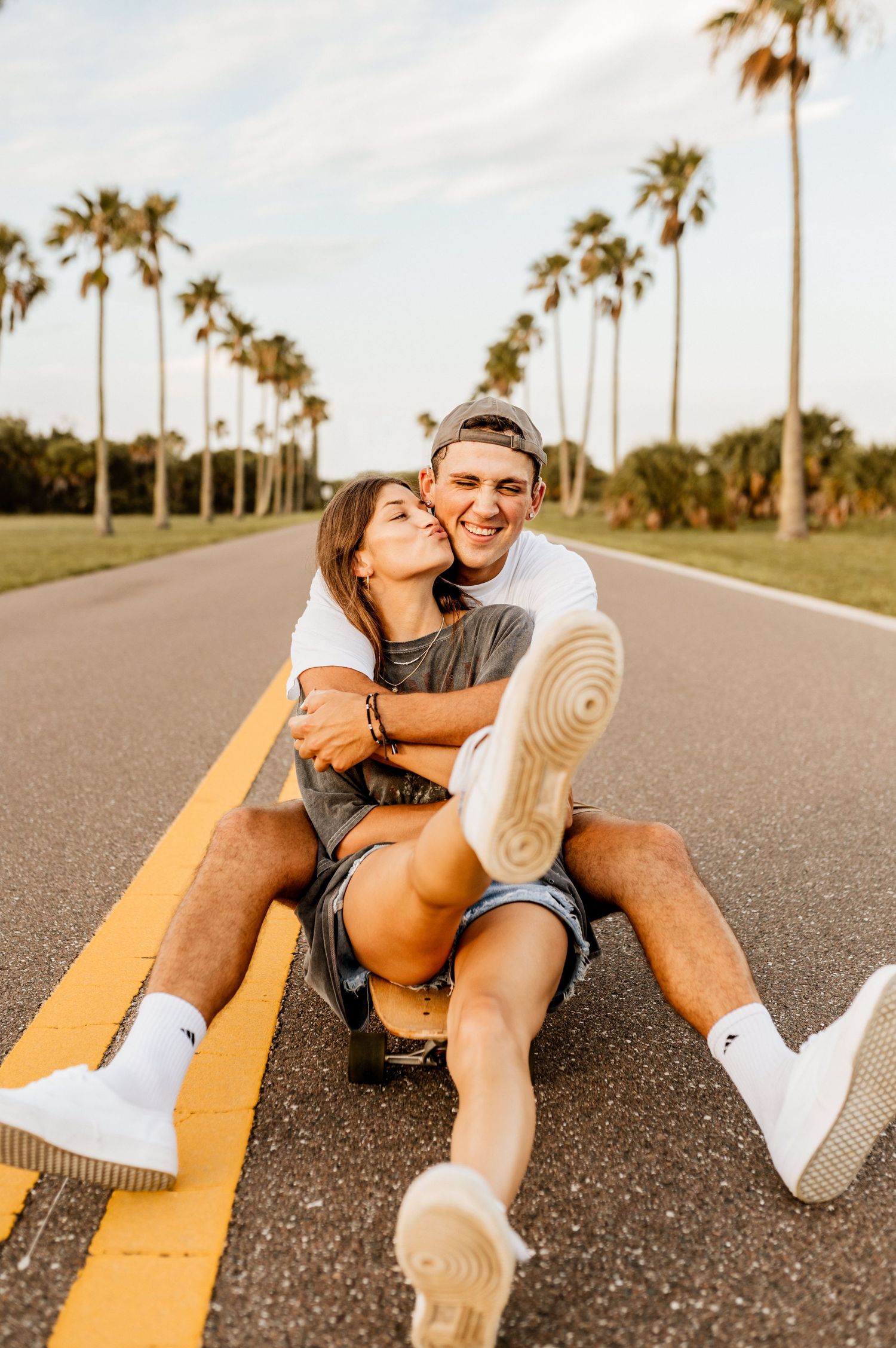 Playful couple sitting together on yellow road line with palm trees in background.