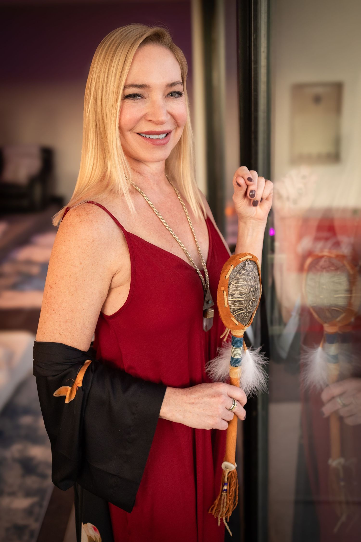 A woman in a burgundy dress and black kimono holds a turtle rattle while smiling in an indoor setting during event photography.