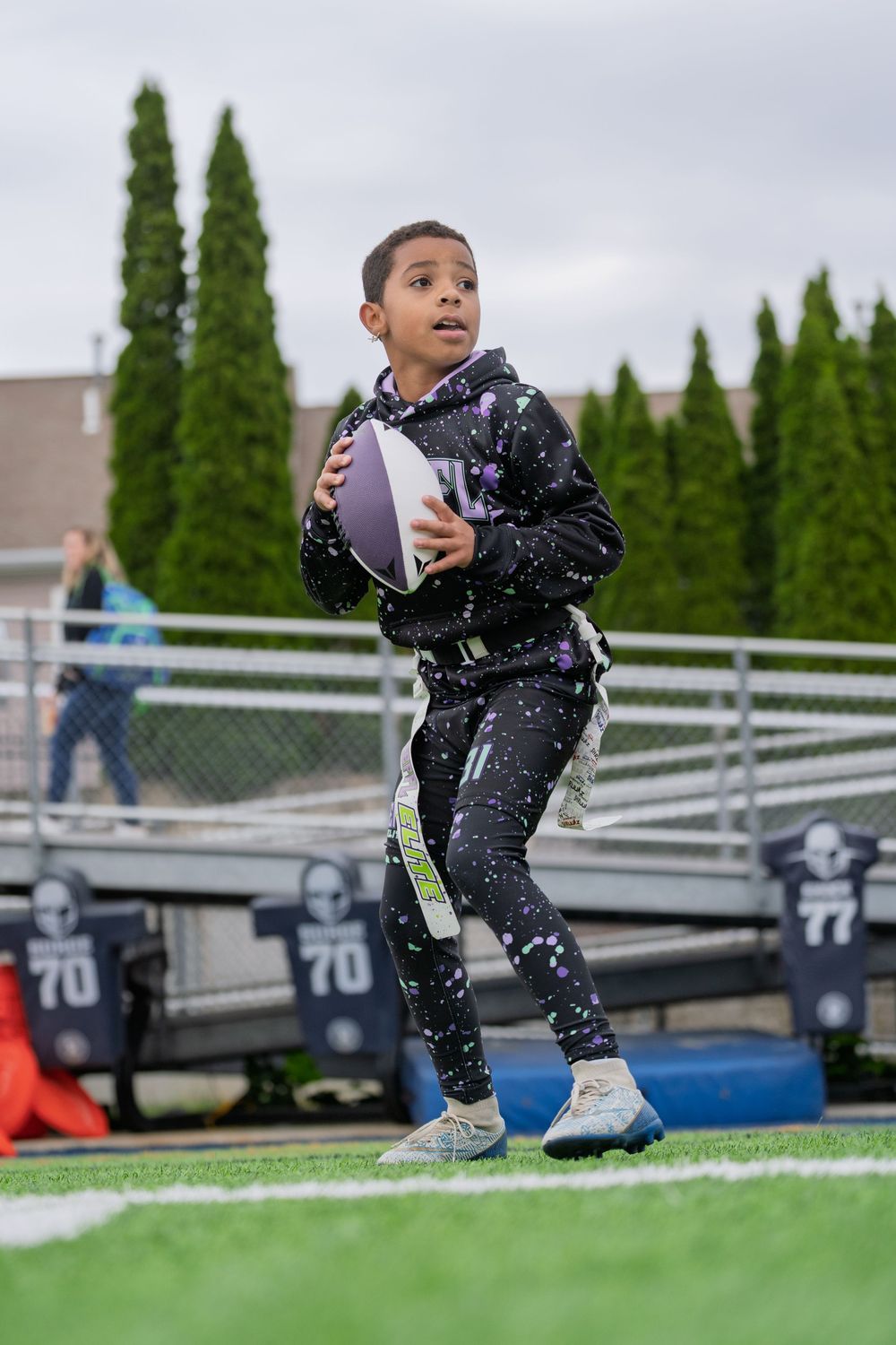 A young athlete in a splatter-patterned tracksuit stands on a sports field with bleachers and cypress trees in the background.