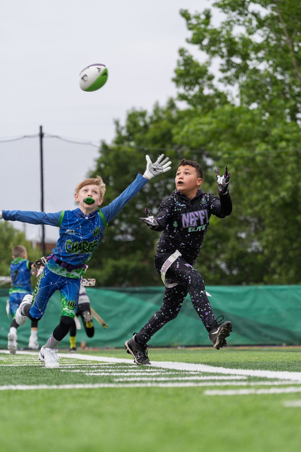 Two young athletes in athletic gear jump to catch a football during practice on a turf field.