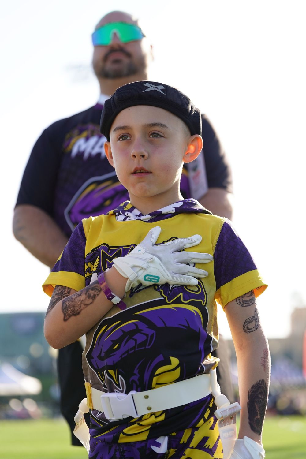 Young baseball player in purple and yellow uniform holds hand up in fielding position on sunny outdoor field.