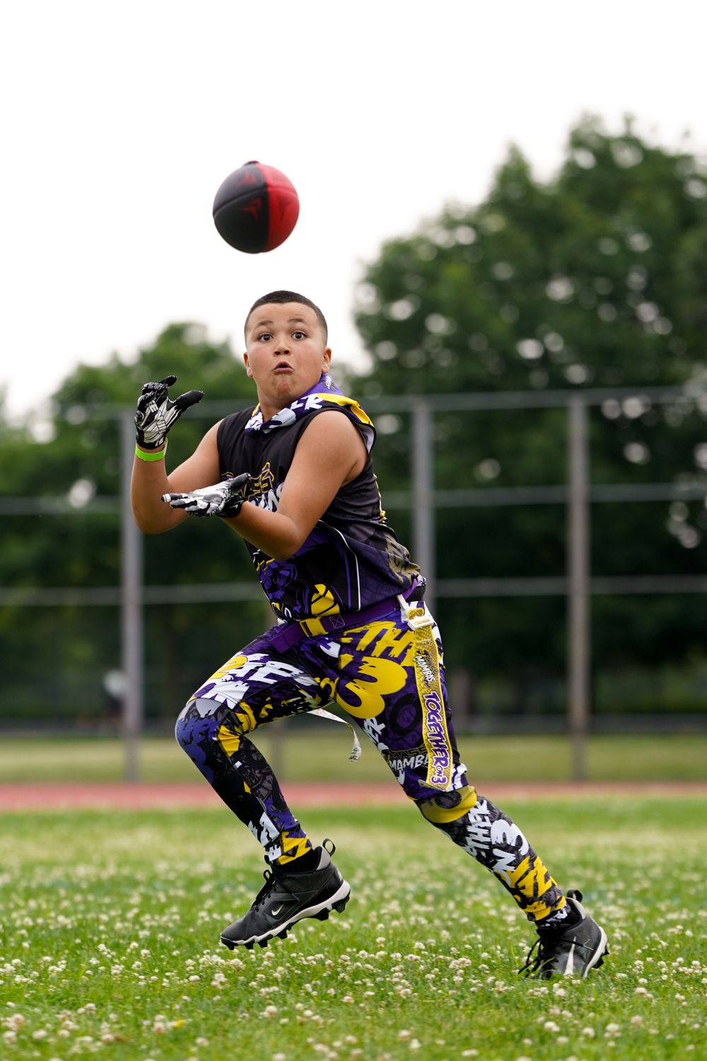 Youth athlete in purple and yellow uniform catches a football while running on a grassy field during practice.