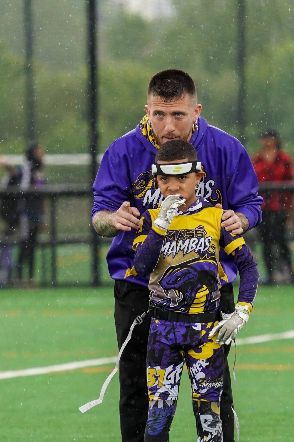 A coach helps adjust gear on a young player wearing a purple and yellow lacrosse uniform on a turf field.