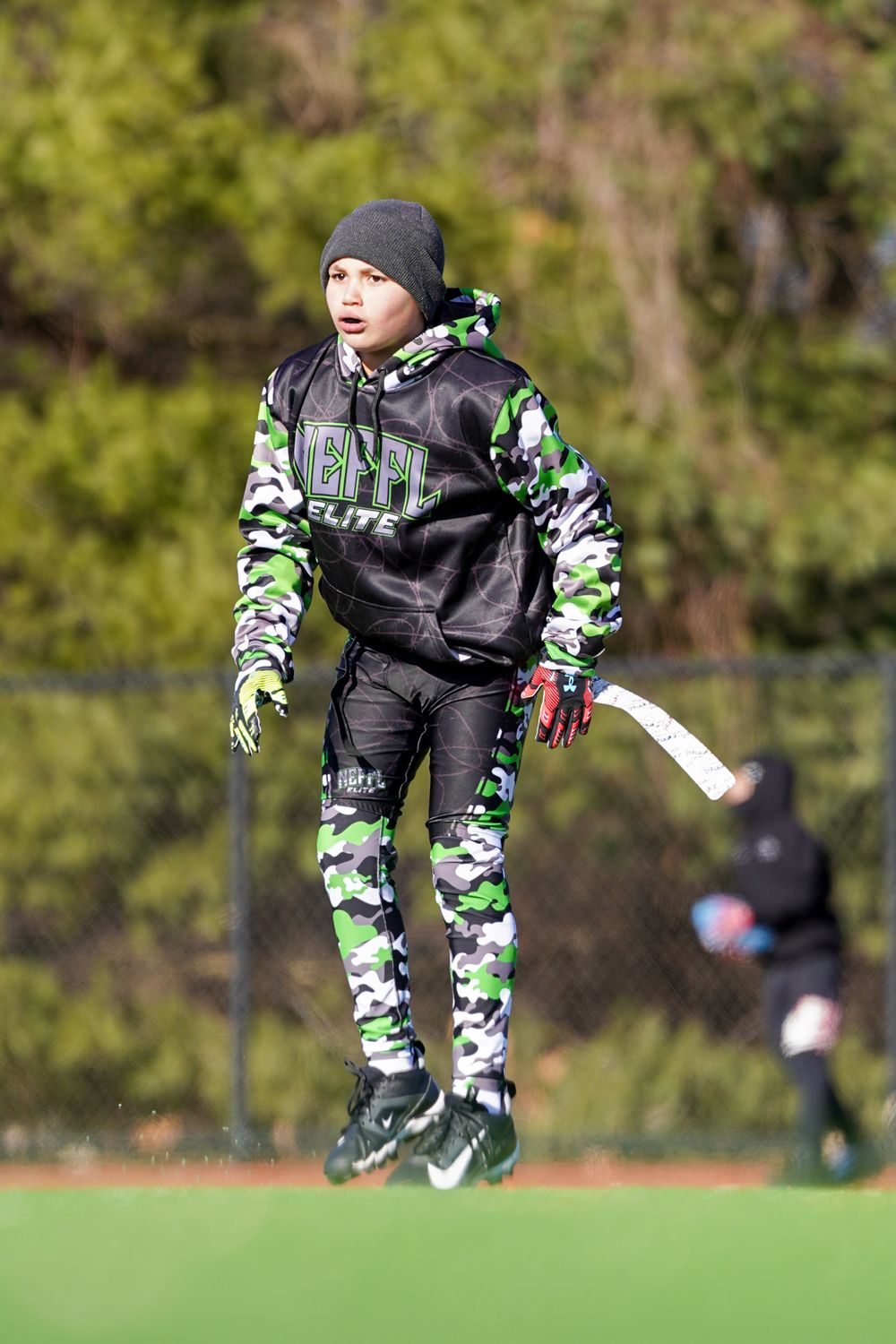 Young field hockey player in camouflage gear and beanie jumps during practice on an outdoor turf field.