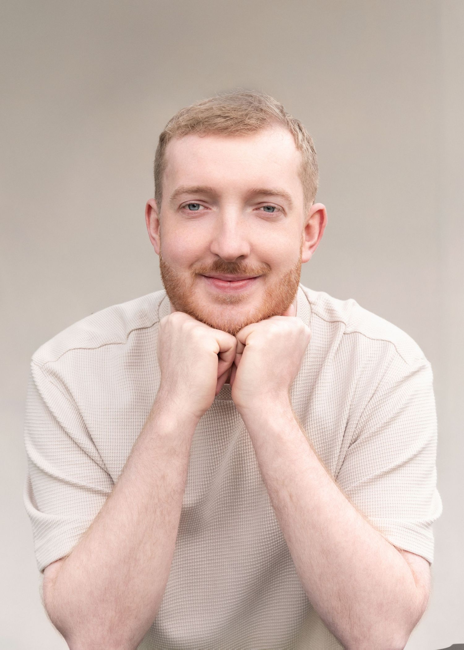 Person wearing a cream colored sweater poses with hands under chin in a gentle portrait against neutral background.