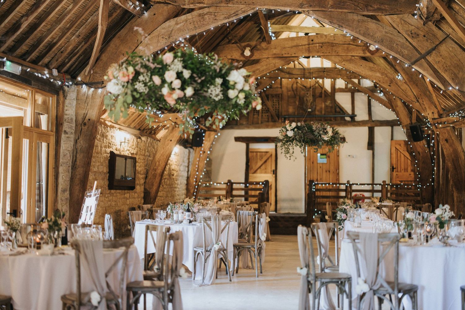 A rustic barn wedding venue with elegant white tables, floral arrangements and exposed wooden beams overhead creating a romantic ambiance.