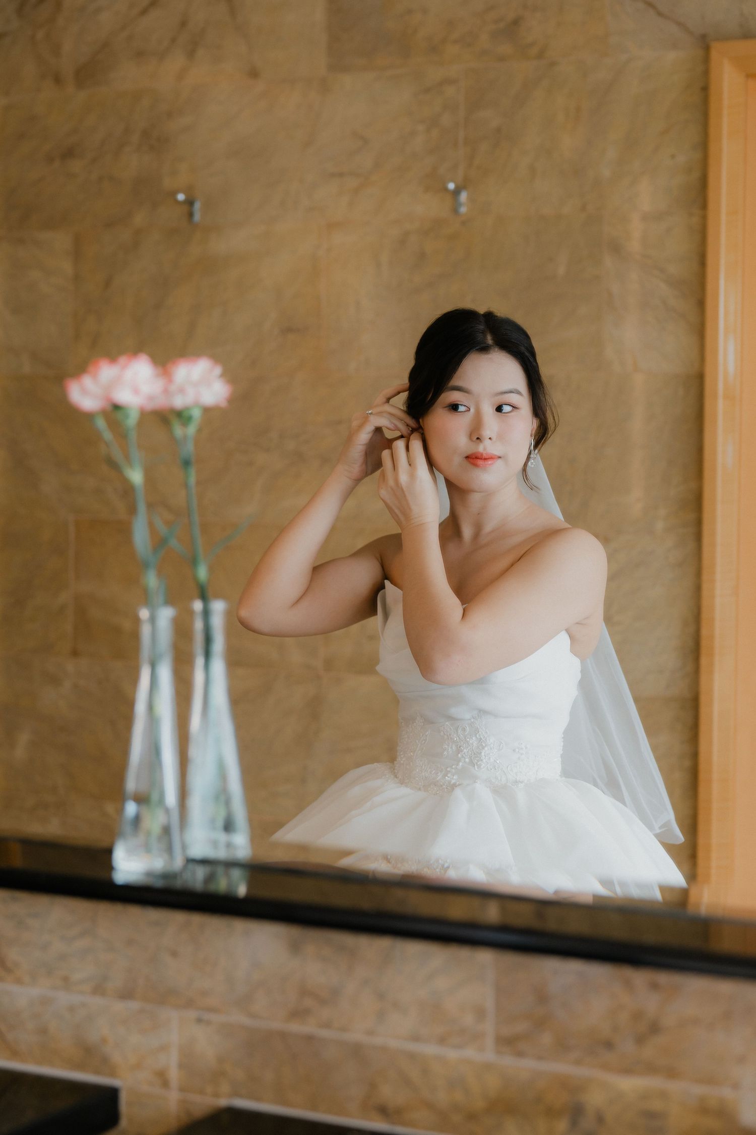 A white wedding dress is reflected in a mirror with pink flowers in a vase on display.