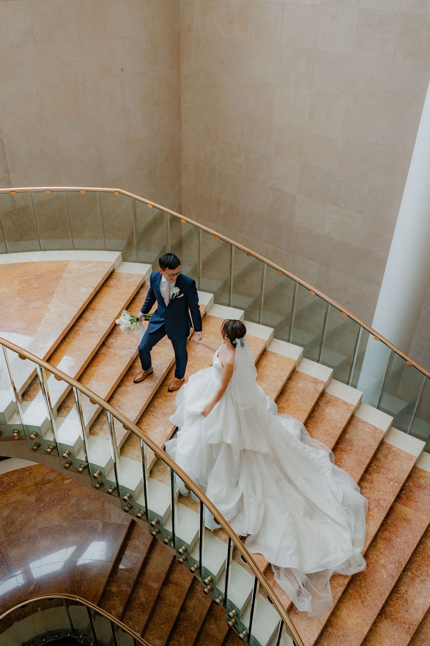 A bride and groom descending an elegant curved marble staircase with a golden handrail in a luxurious venue.