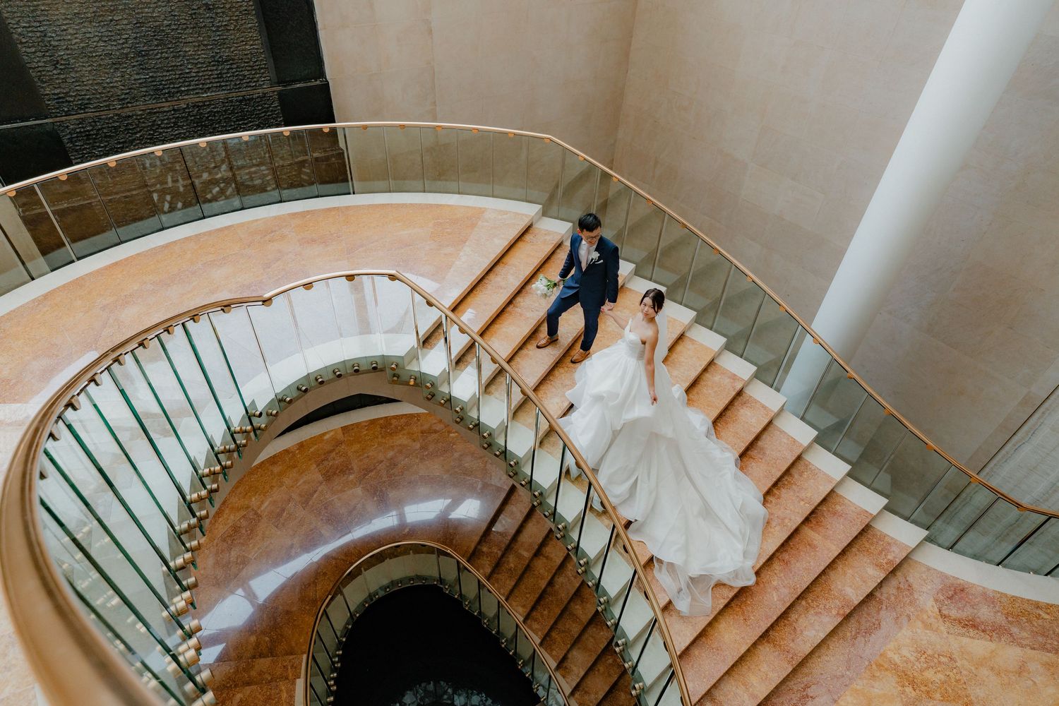 A couple in wedding attire descends an elegant spiral staircase with white marble steps and metal railings.