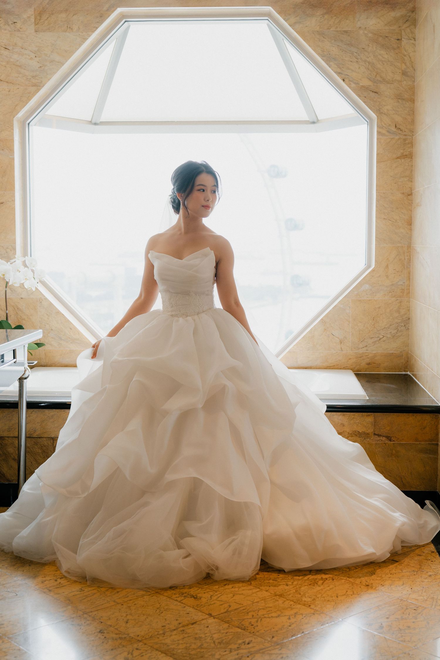 A bride poses in an elegant strapless wedding gown with layered tulle skirt by an octagonal window.