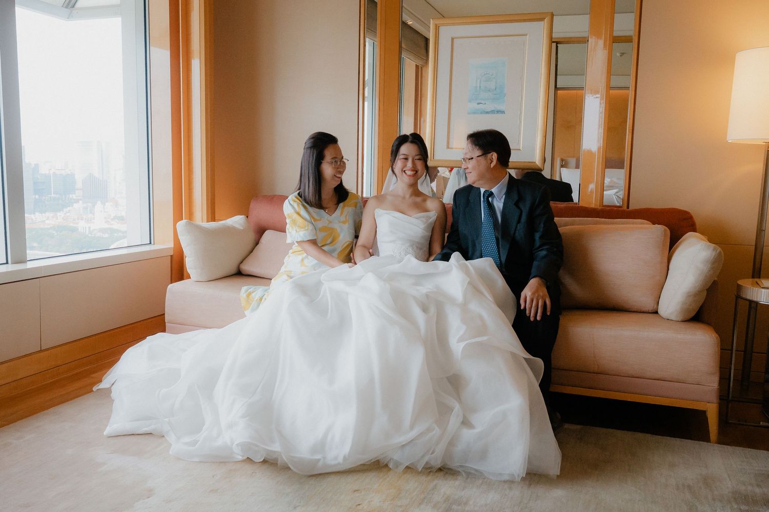 Wedding party relaxing on a beige couch in a sunlit hotel room.