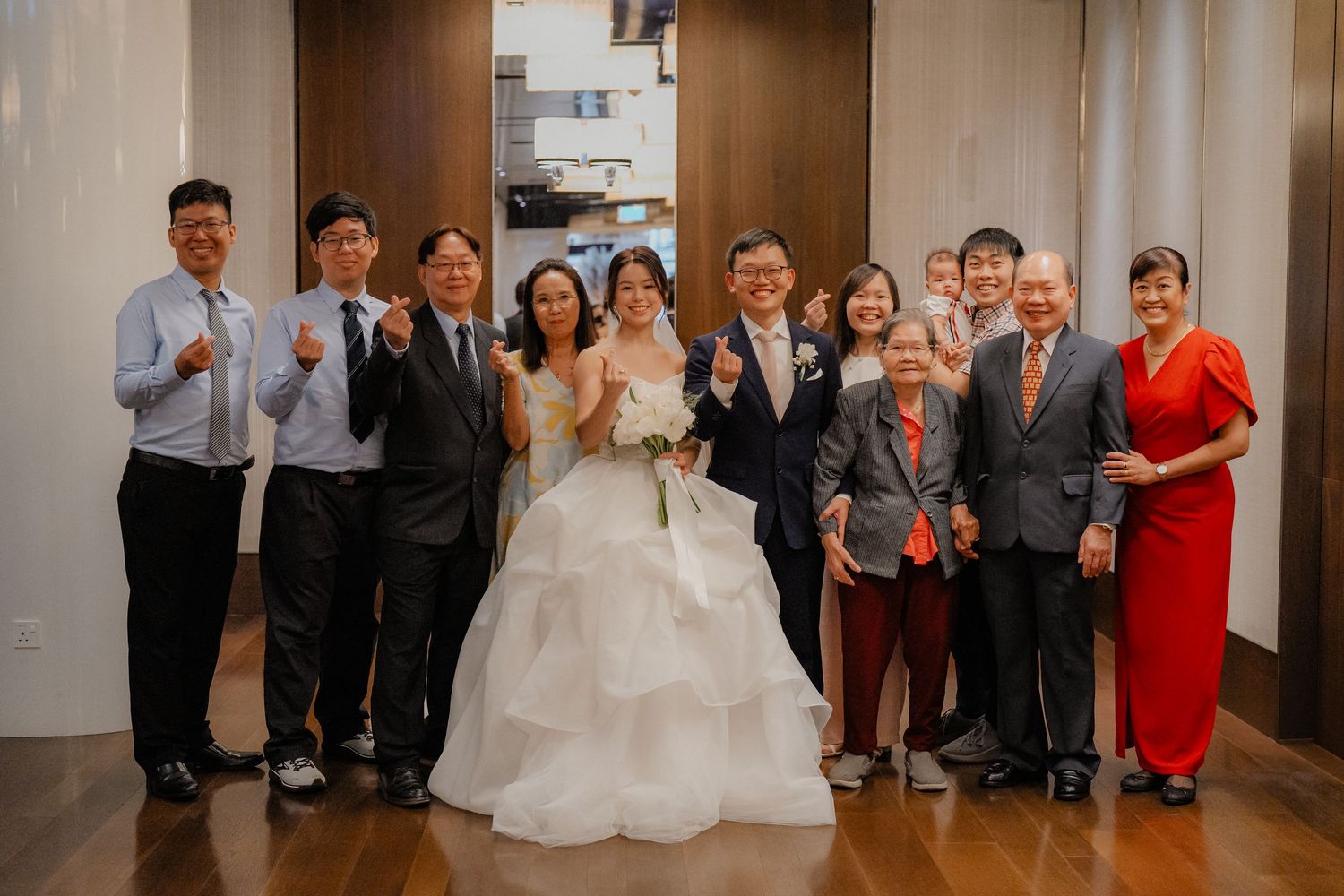 A wedding party group poses together in a formal indoor setting with people wearing suits and a flowing white gown.