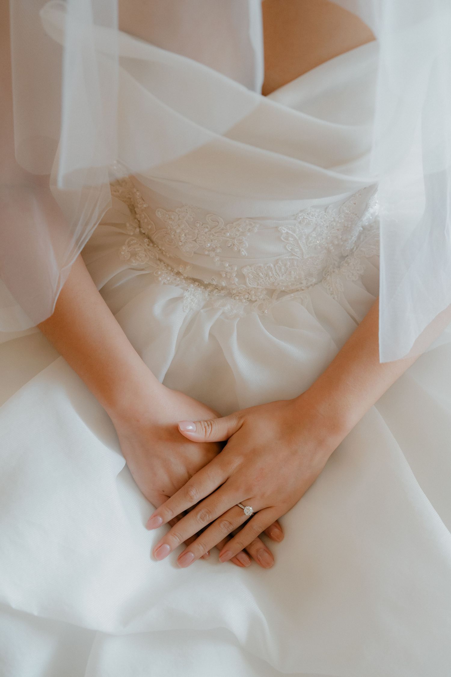 Close-up of folded hands resting on a white wedding dress with delicate lace details.