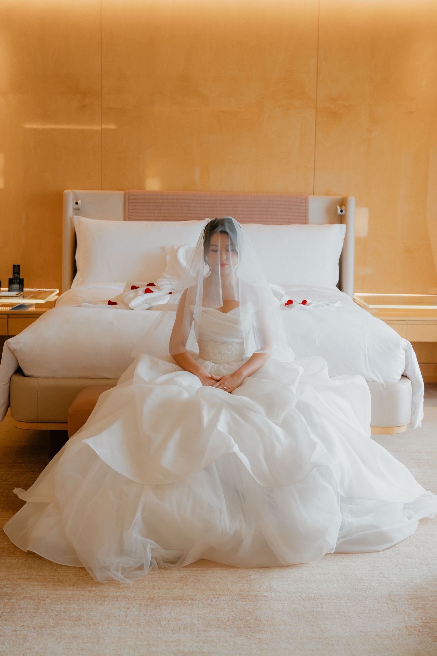 A bride in a flowing white wedding gown and veil sits serenely on a hotel bed with rose petals scattered around.