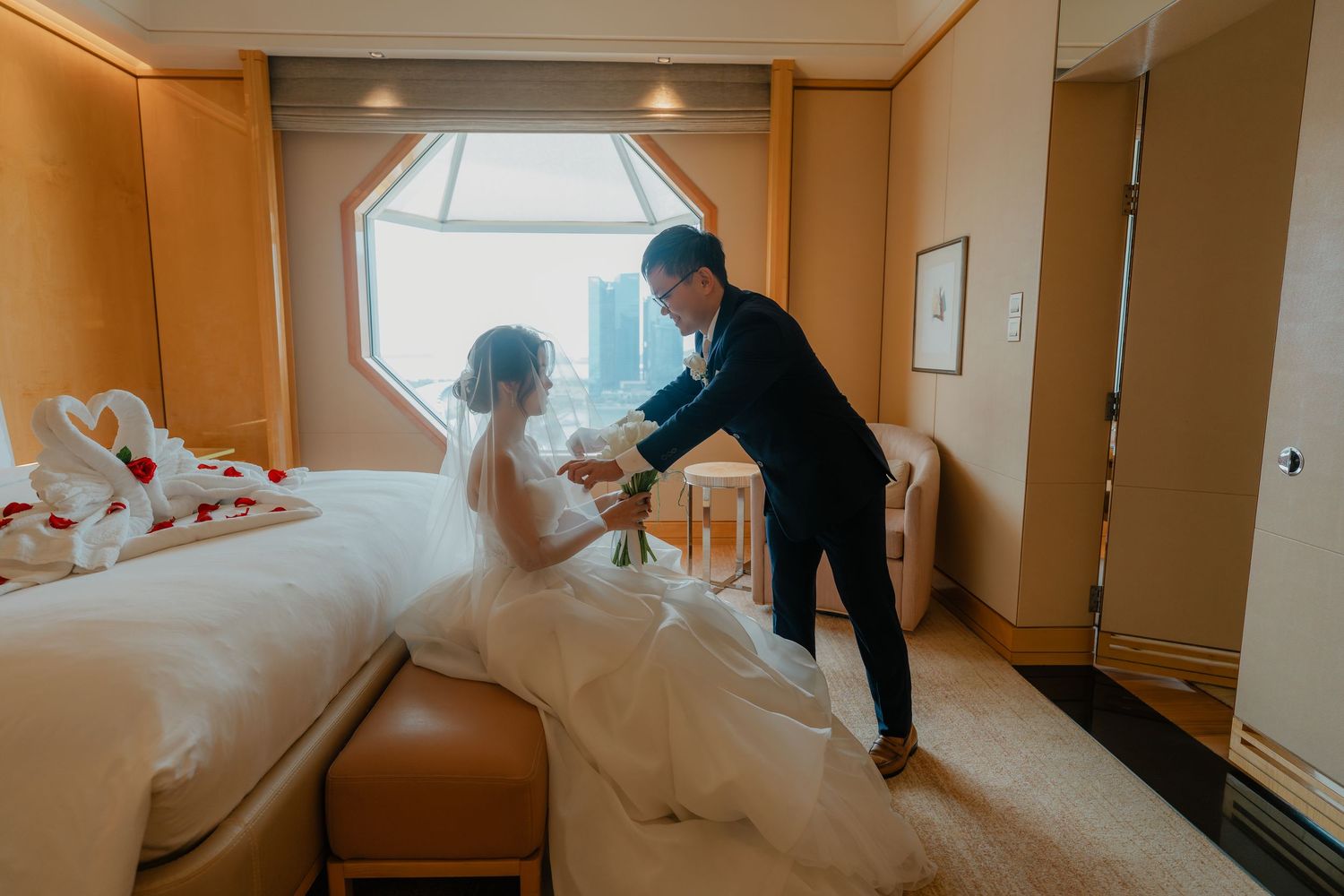 A couple in wedding attire prepare in an elegant hotel room with an octagonal window.