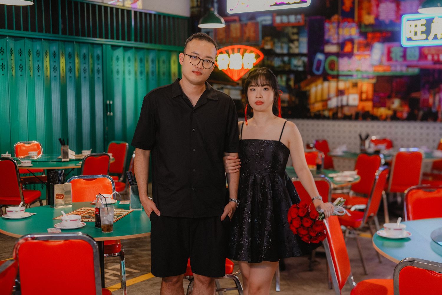 A couple in matching black outfits poses in a retro-style Asian diner with neon signs.