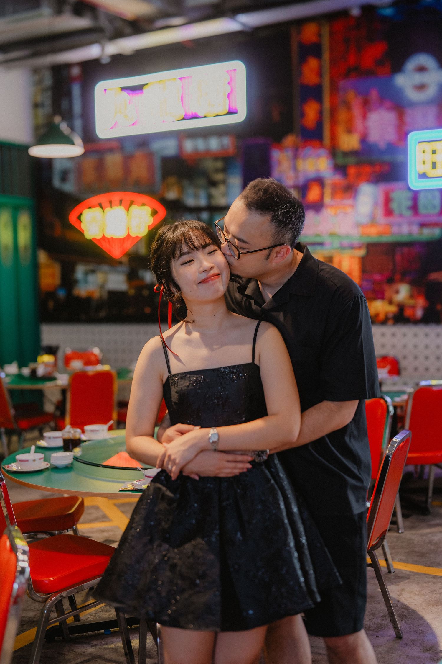 A couple embracing in a dimly lit restaurant with vibrant neon signs illuminating the scene.