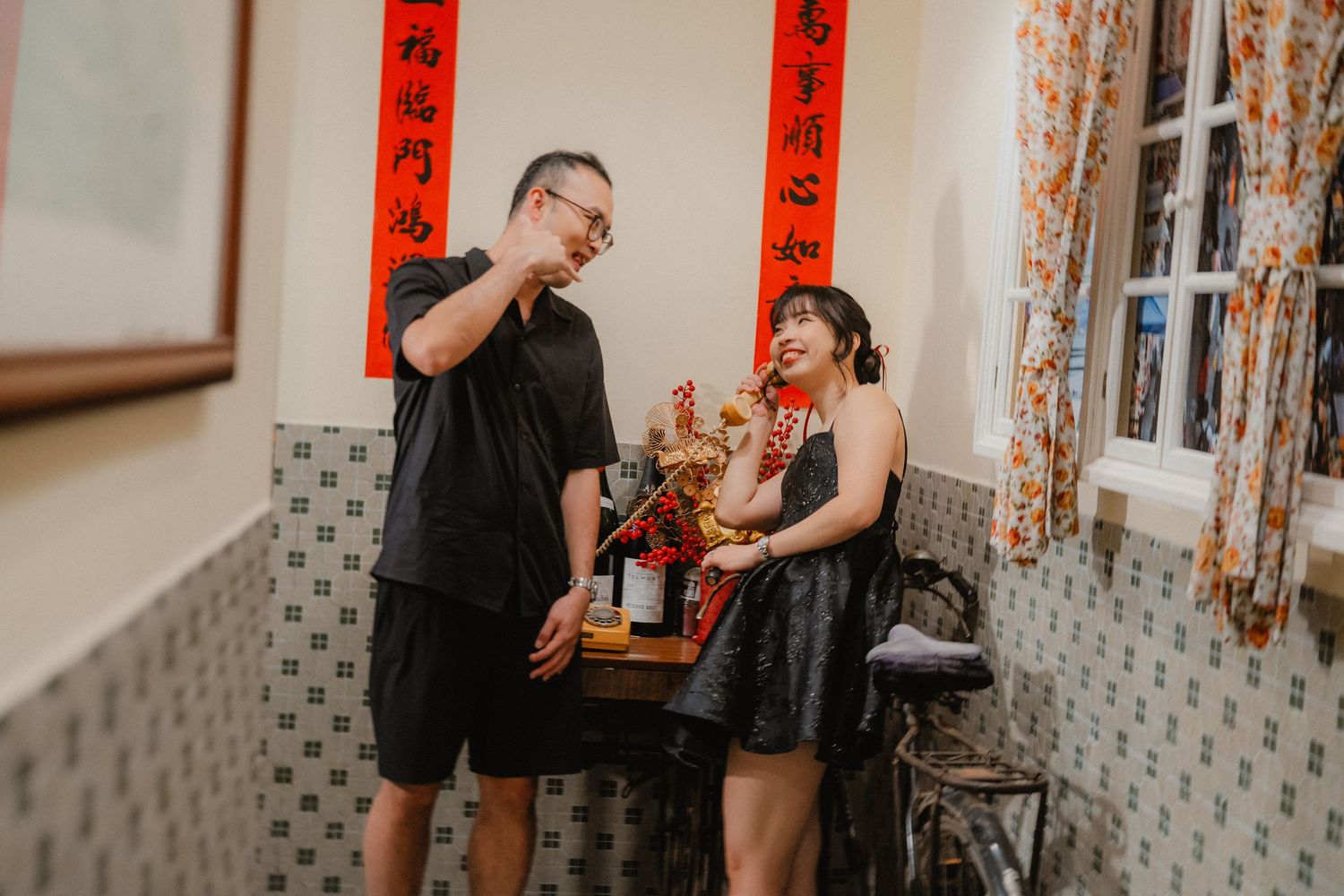 Couple in black outfits sharing drinks while celebrating Chinese New Year with red decorations in the background.