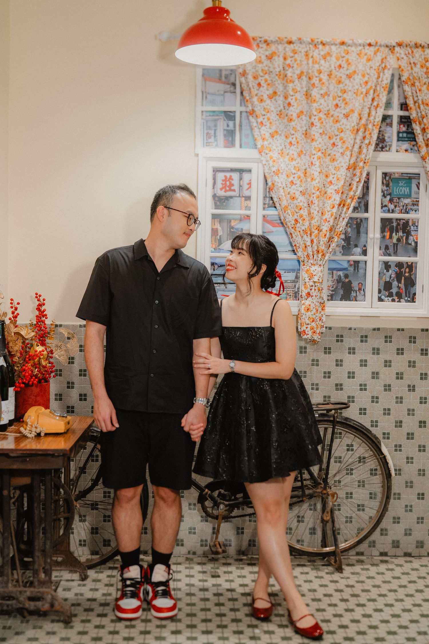 A couple in coordinated black outfits poses in a vintage-style room with floral curtains and a bicycle.