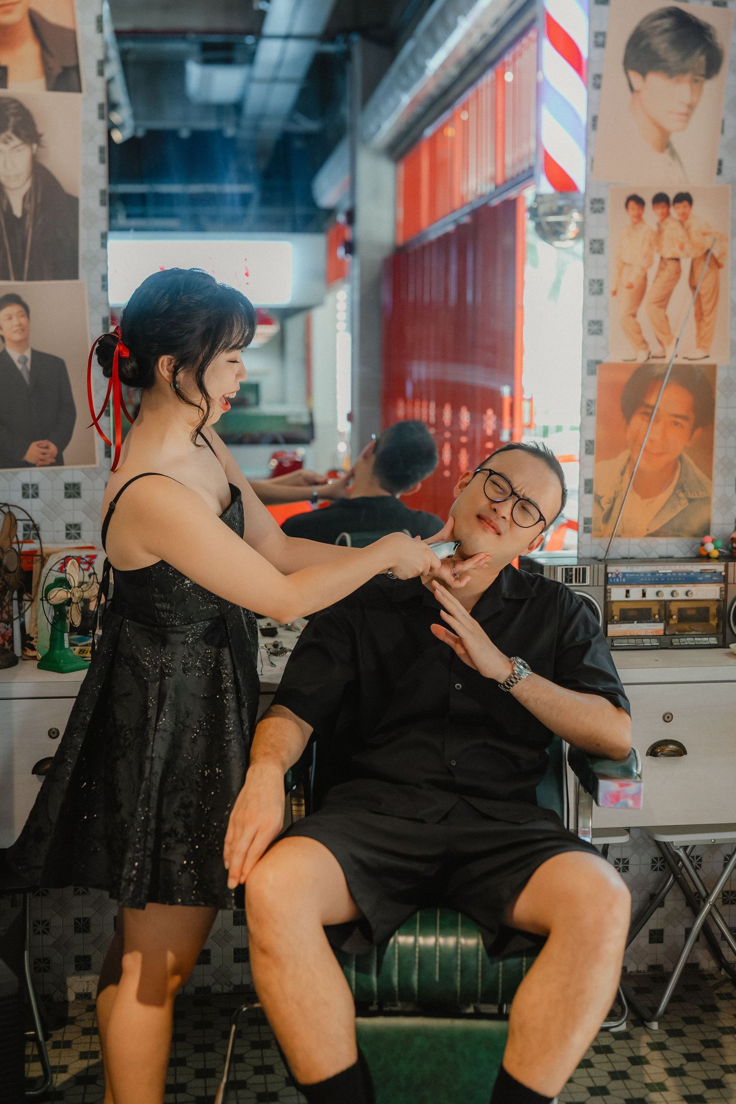 A couple playfully poses at a retro-style barbershop with vintage posters on the wall.