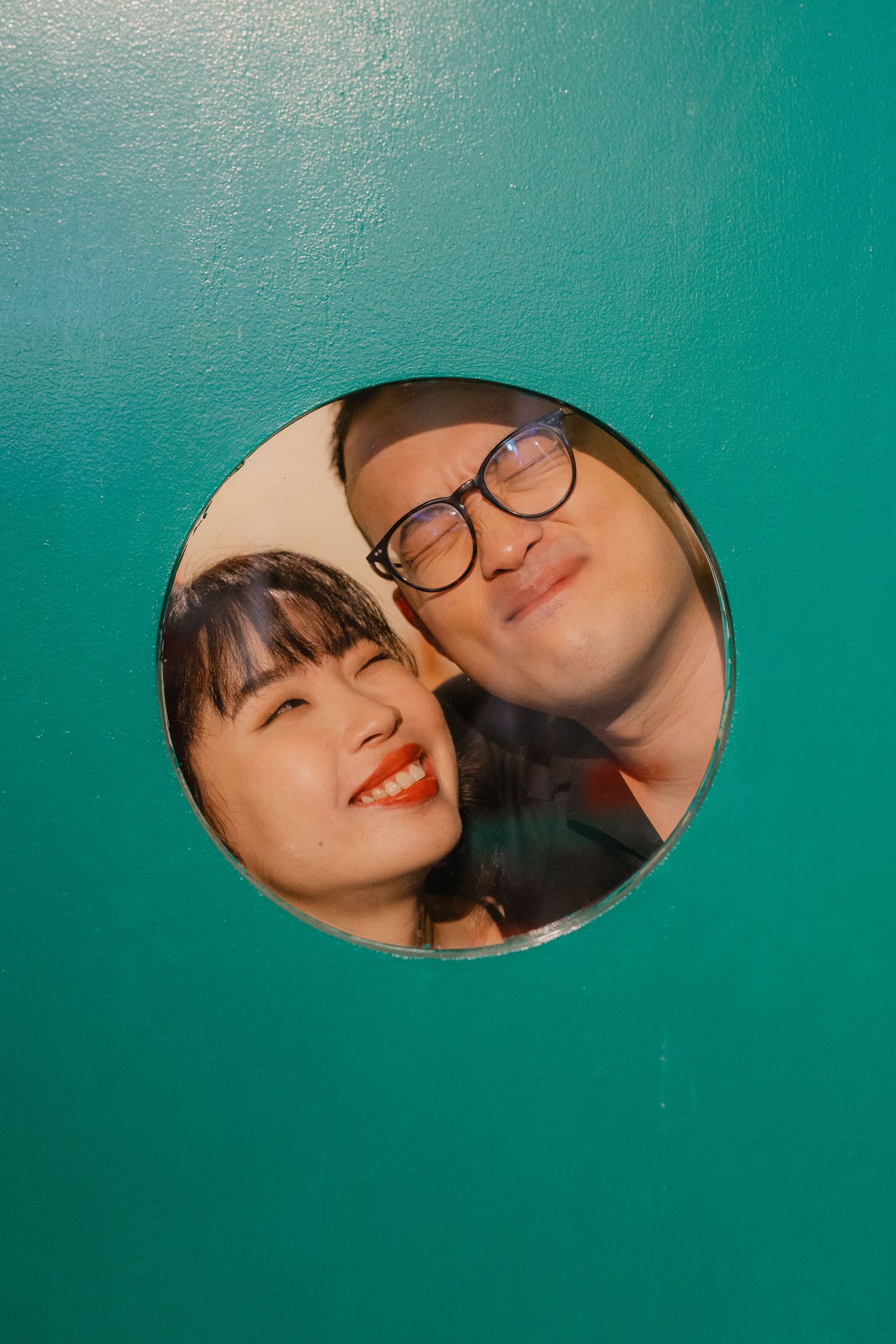 A couple smiles through a circular window frame against a turquoise background.