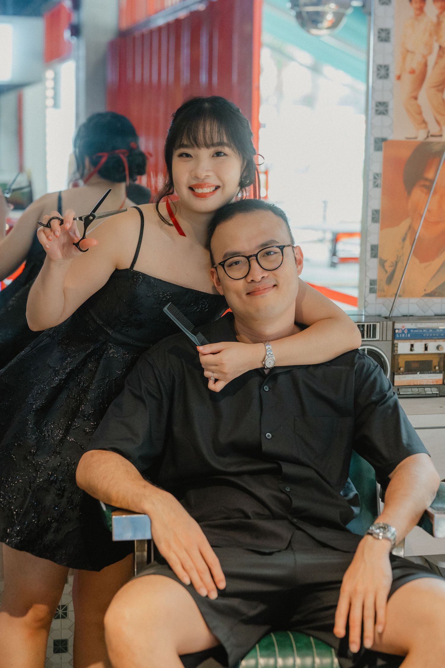 A couple poses together at a colorful storefront, with one person wearing a black dress and another in dark casual attire.