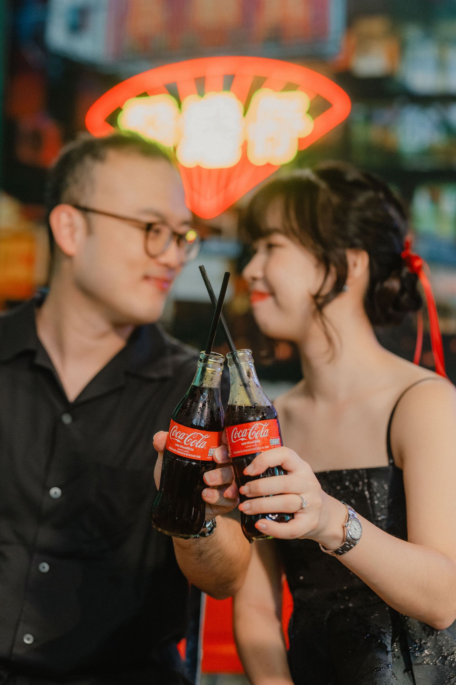A couple toasting with Coca-Cola bottles under a neon light in an intimate setting.