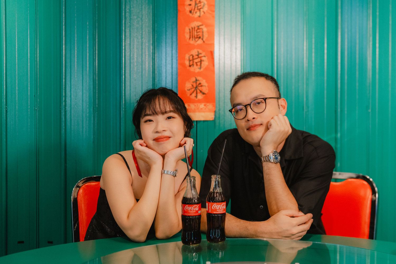 A couple sits at a table with Coca-Cola bottles in front of a turquoise wall.