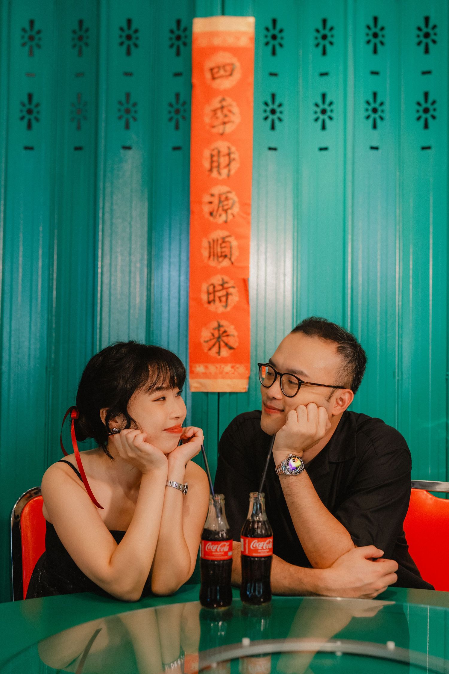 A couple sharing a moment at a table with Coca-Cola bottles against a turquoise wall with Chinese characters.