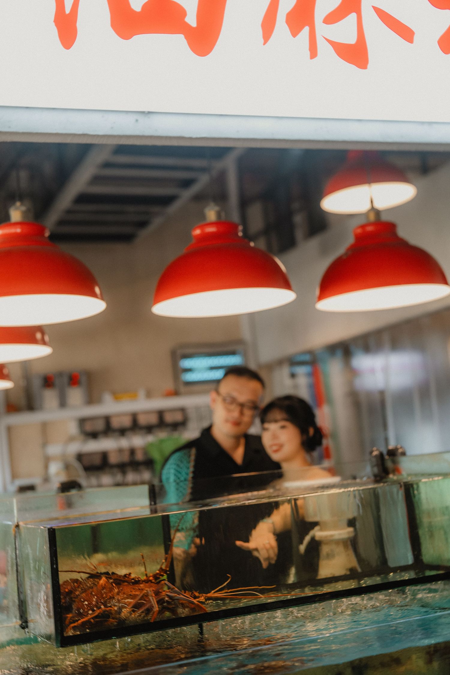 A couple looks at seafood in an aquarium tank under red pendant lamps at an Asian restaurant.