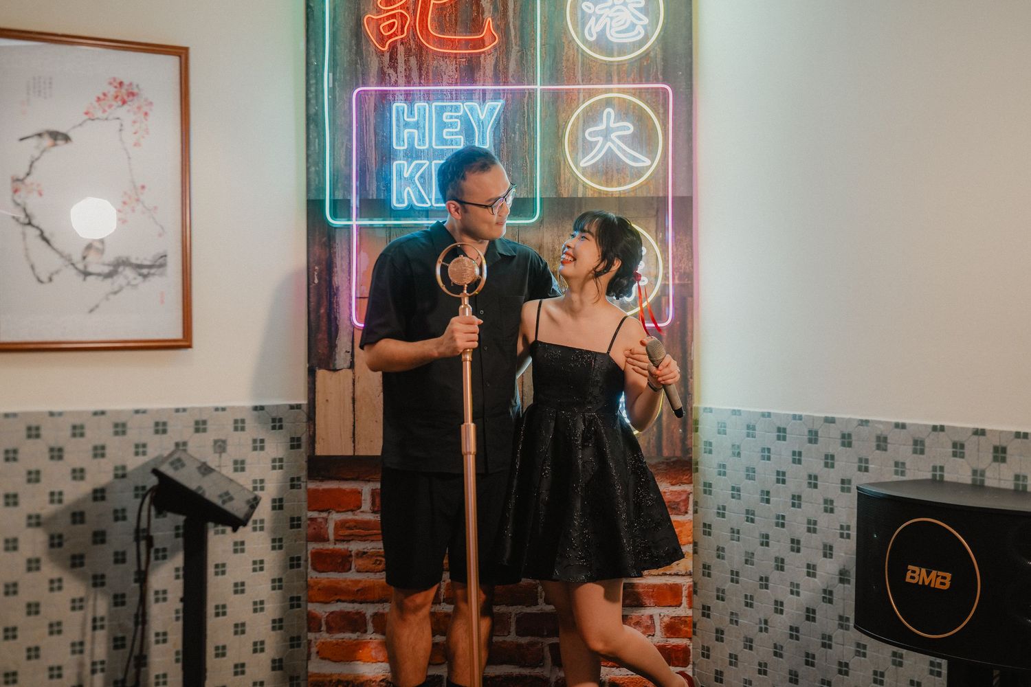 Two performers share a microphone on stage with colorful neon signs glowing in the background at a comedy club.