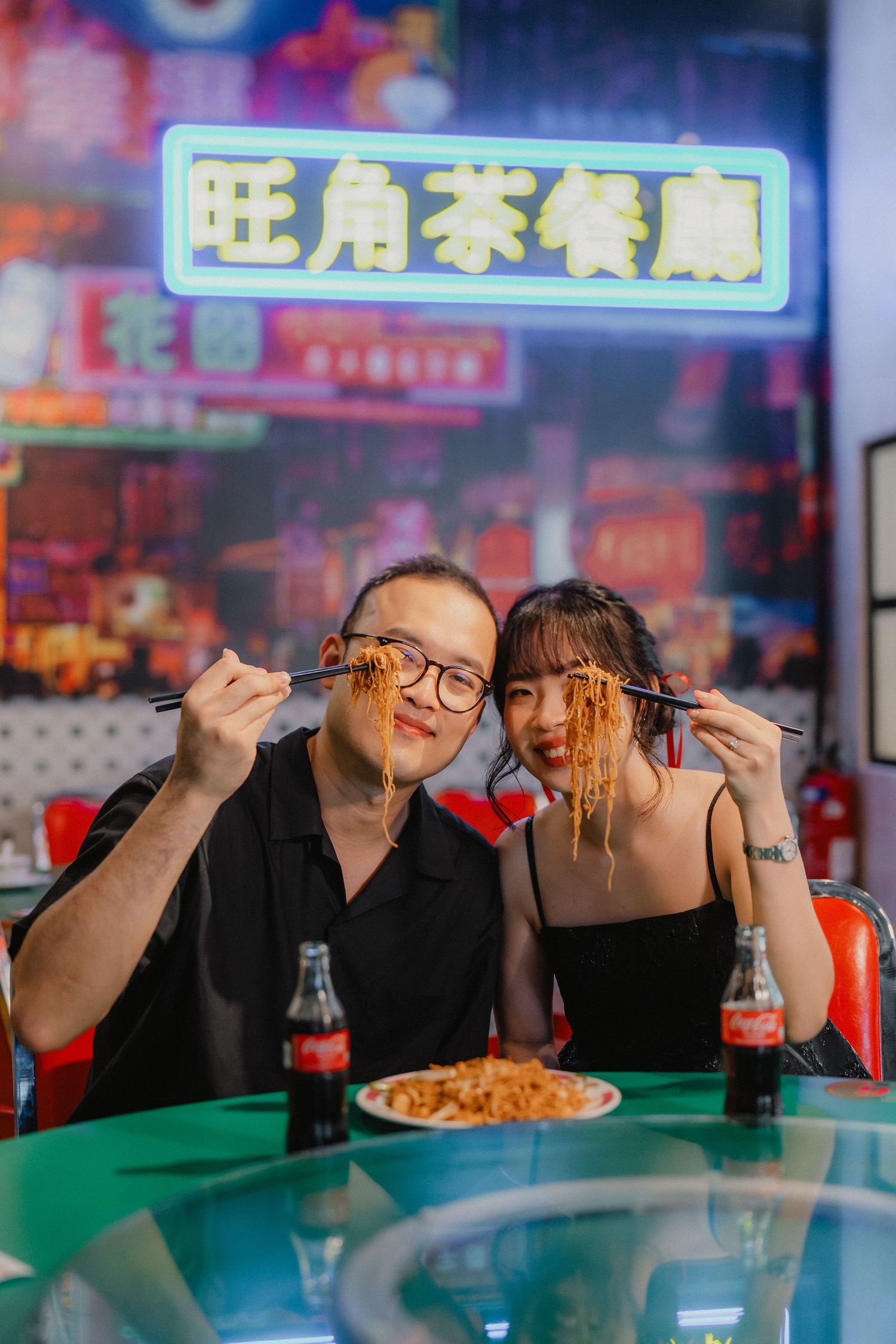 A couple enjoying noodles at a restaurant with neon signs and Coca-Cola bottles on the table.