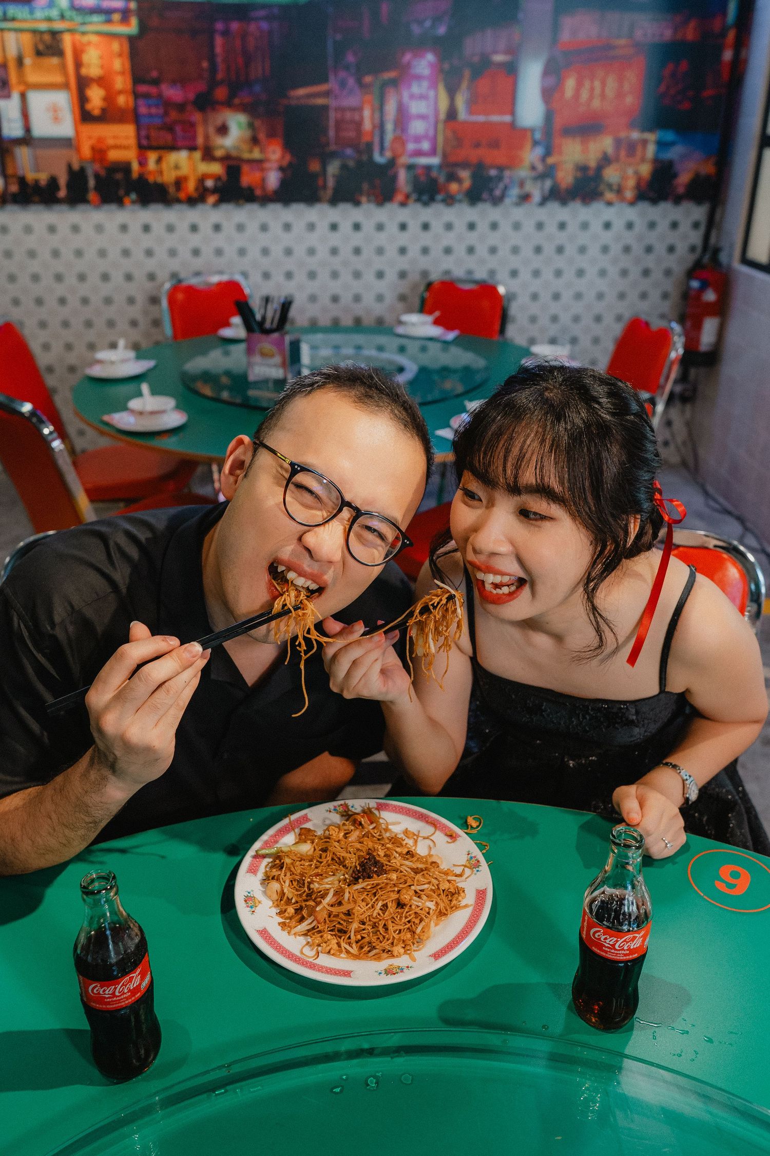 A couple enjoys eating noodles together at a retro-style diner with Coca-Cola bottles and red chairs.
