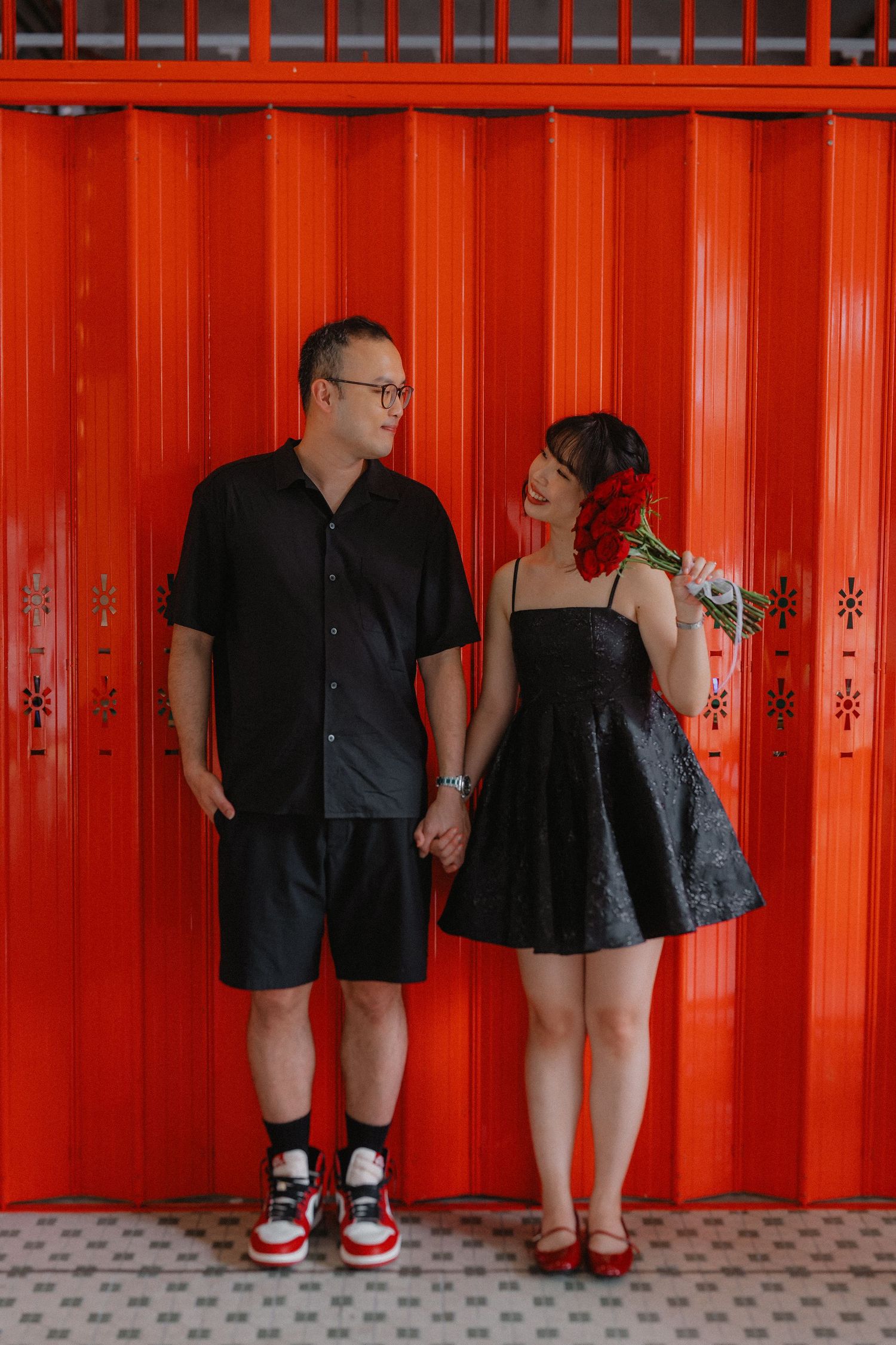 A couple in black outfits posing against a vibrant red wall in a casual setting.