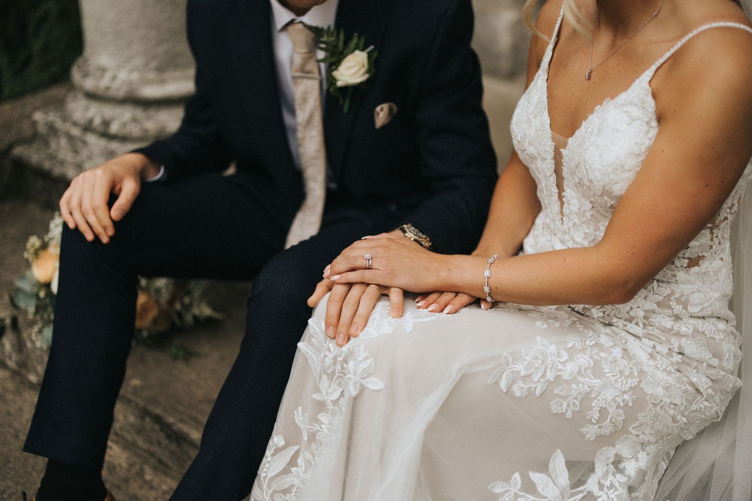 Close-up view of clasped hands with wedding rings against delicate white lace fabric in natural lighting.