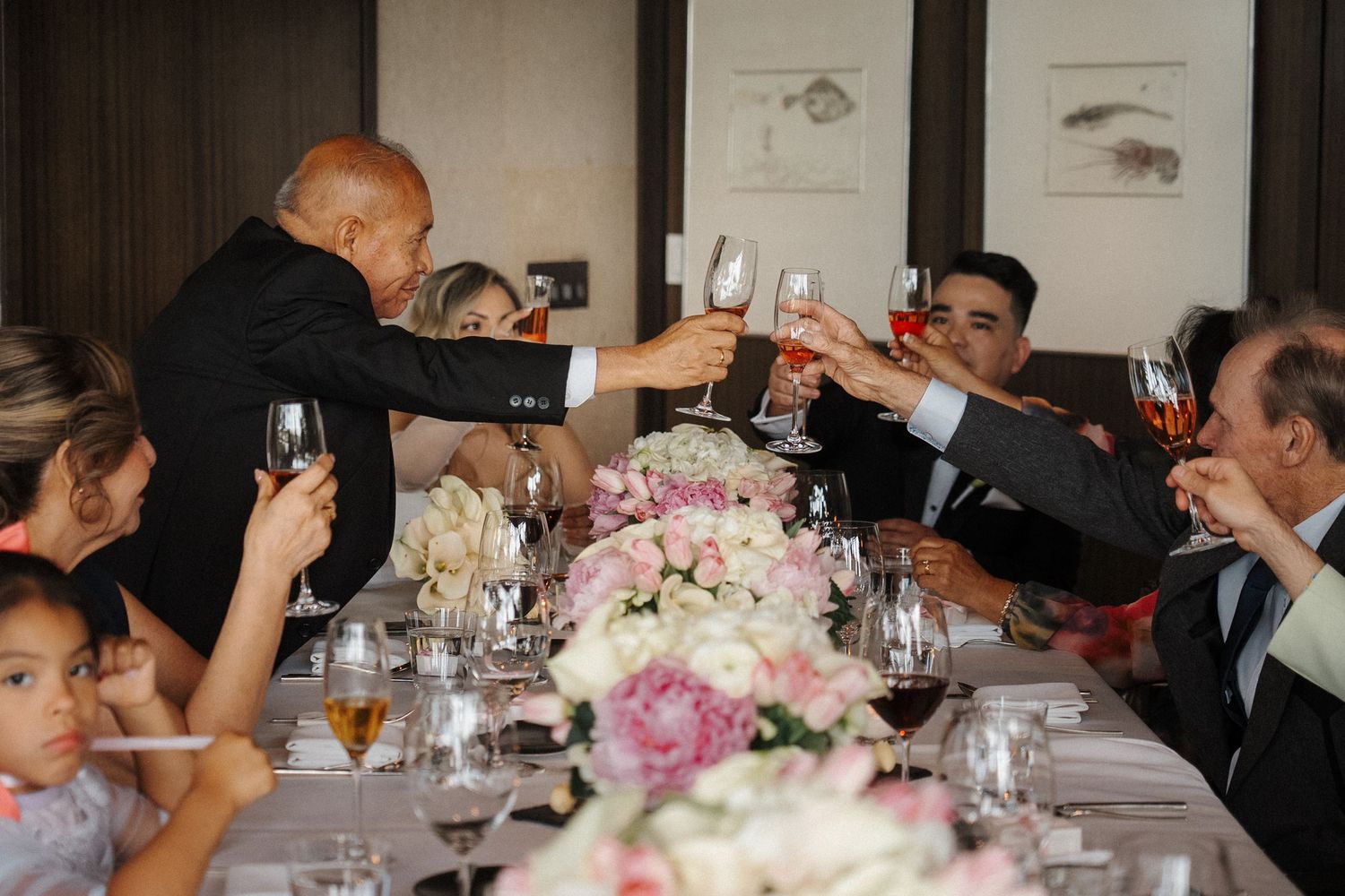 Wedding guests raise wine glasses in a celebratory toast at an elegant dinner table decorated with pink floral centerpieces.
