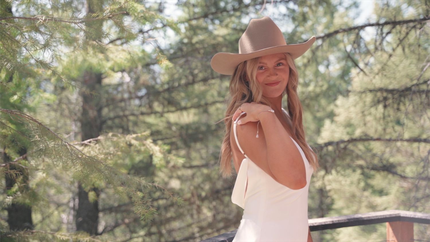 A person in a brown hat and white dress poses gracefully amid tall pine trees and sunlight in an outdoor photoshoot.