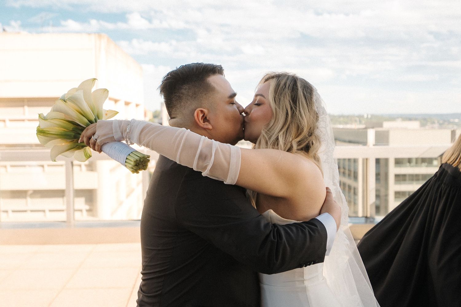 Two people embrace and kiss in romantic poses while standing together in front of a light colored background.