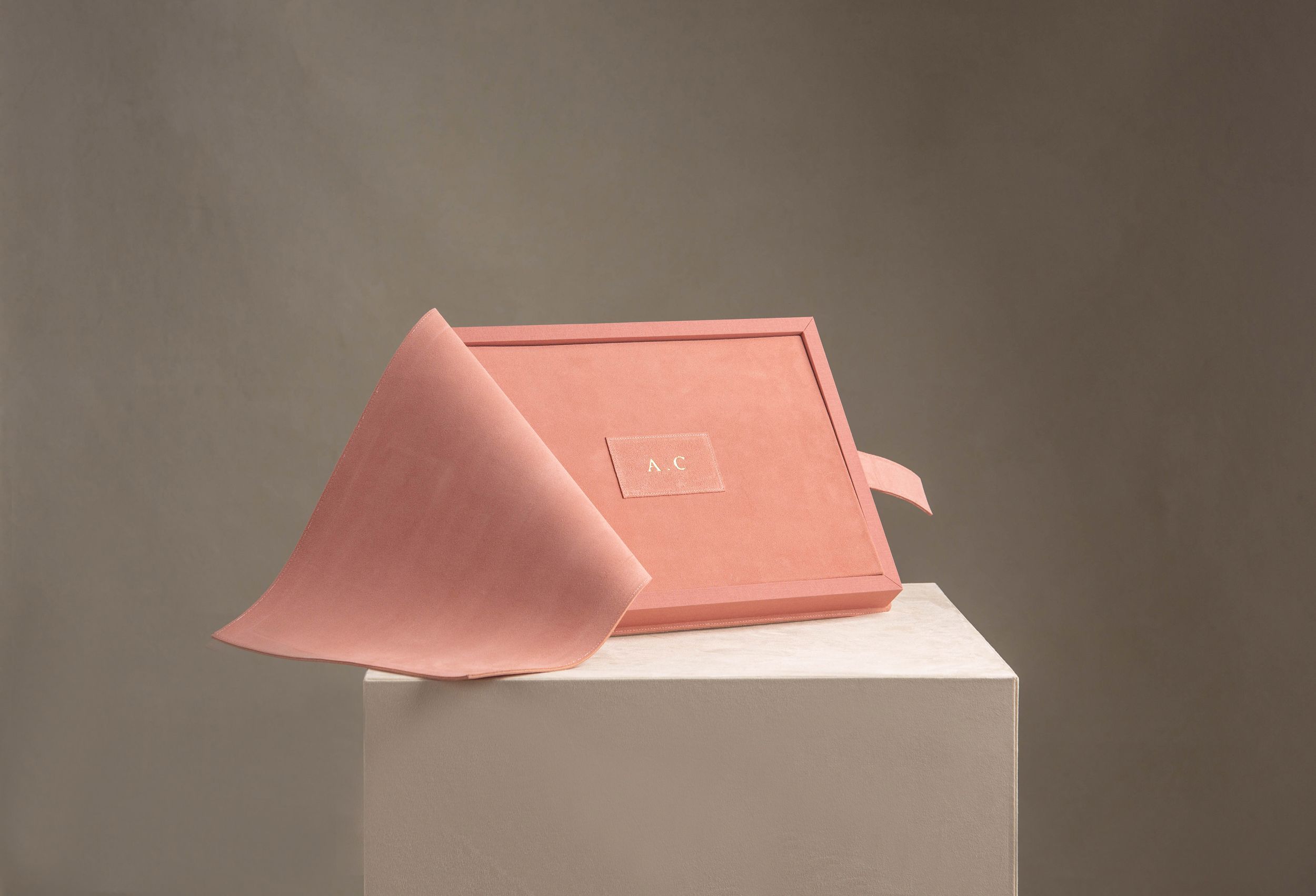 A soft pink folded rectangular bar of soap rests on a white pedestal against a muted brown background.